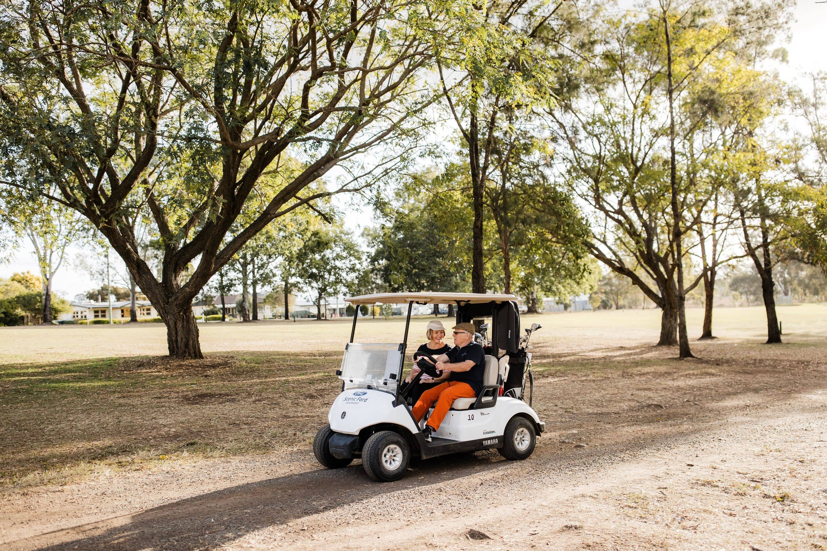 Two residents in a white golf cart on a dirt path, with golf clubs in the back. Trees and grassy grounds define the community setting.