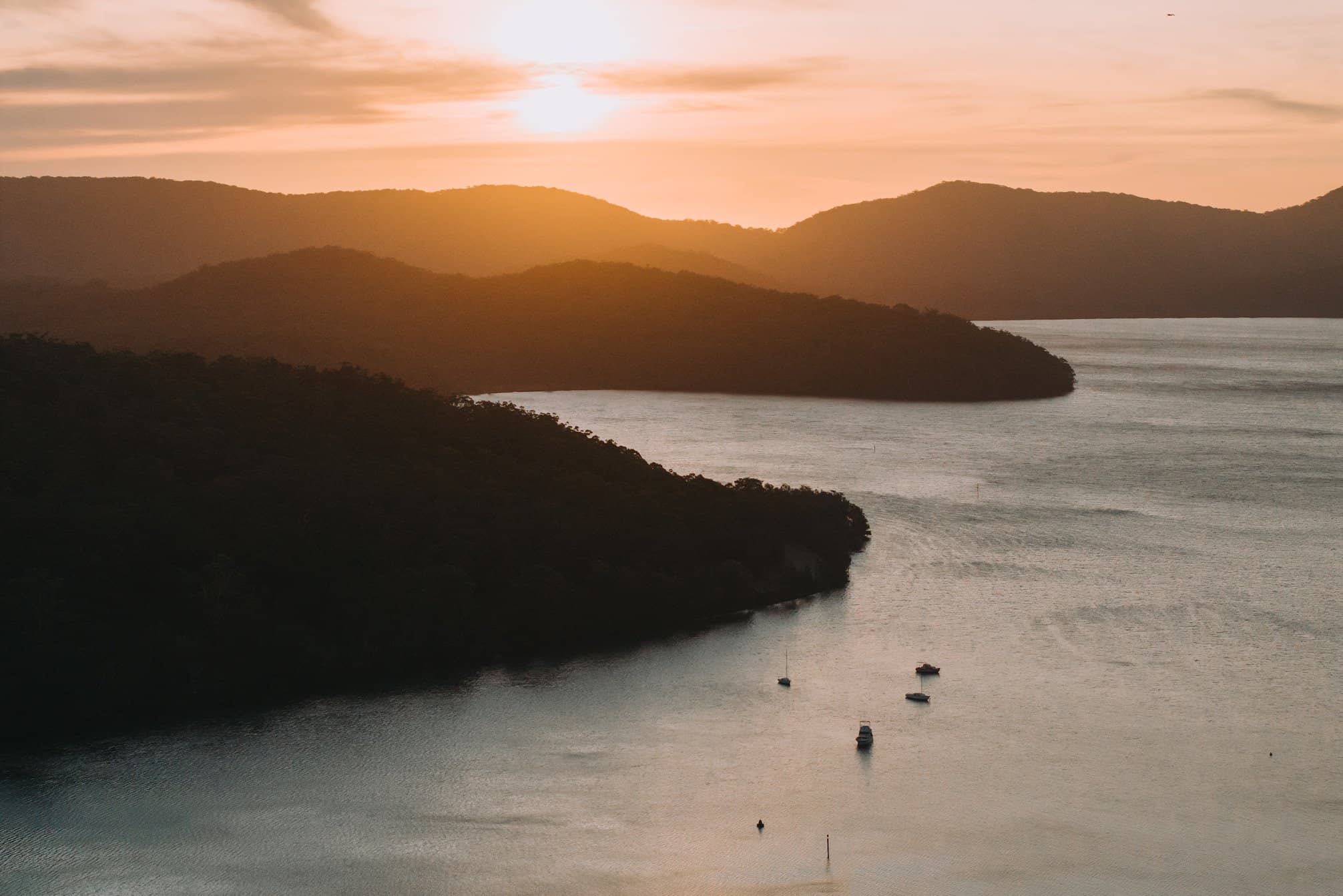 Sunset over a tranquil bay with forested hills and several boats on the water.