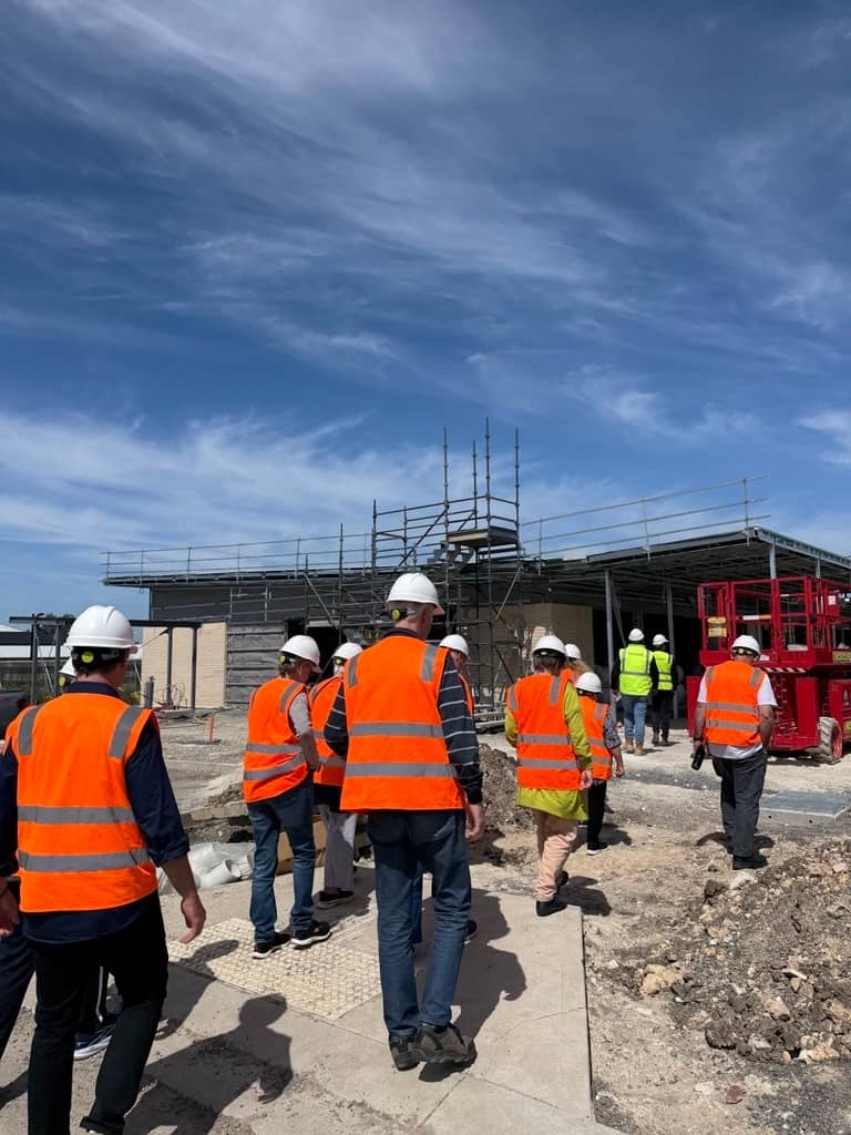 A group of people in hard hats and orange safety vests walk past a construction site with scaffolding.