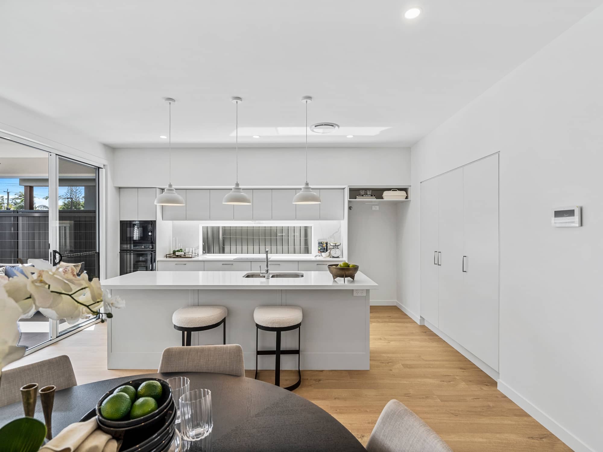Bright, modern kitchen in an Ingenia Lifestyle home with light grey cabinetry, island seating, and dining area. Sliding doors lead outside.