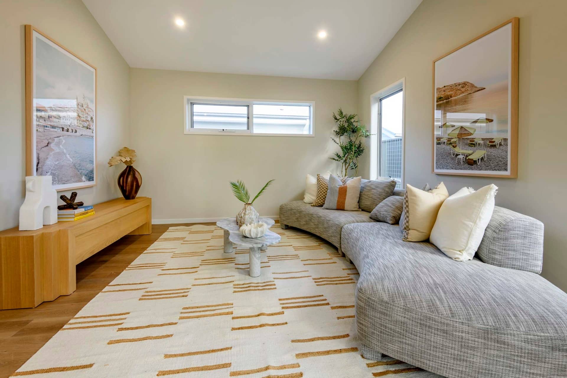 Curved grey sofa with decorative pillows in a living room with two framed beach scenes and a wooden console table.