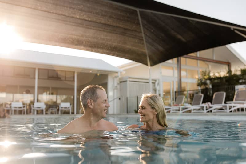 A man and a woman swim in a pool at an Ingenia Lifestyle community, with lounge chairs and a building in the background.