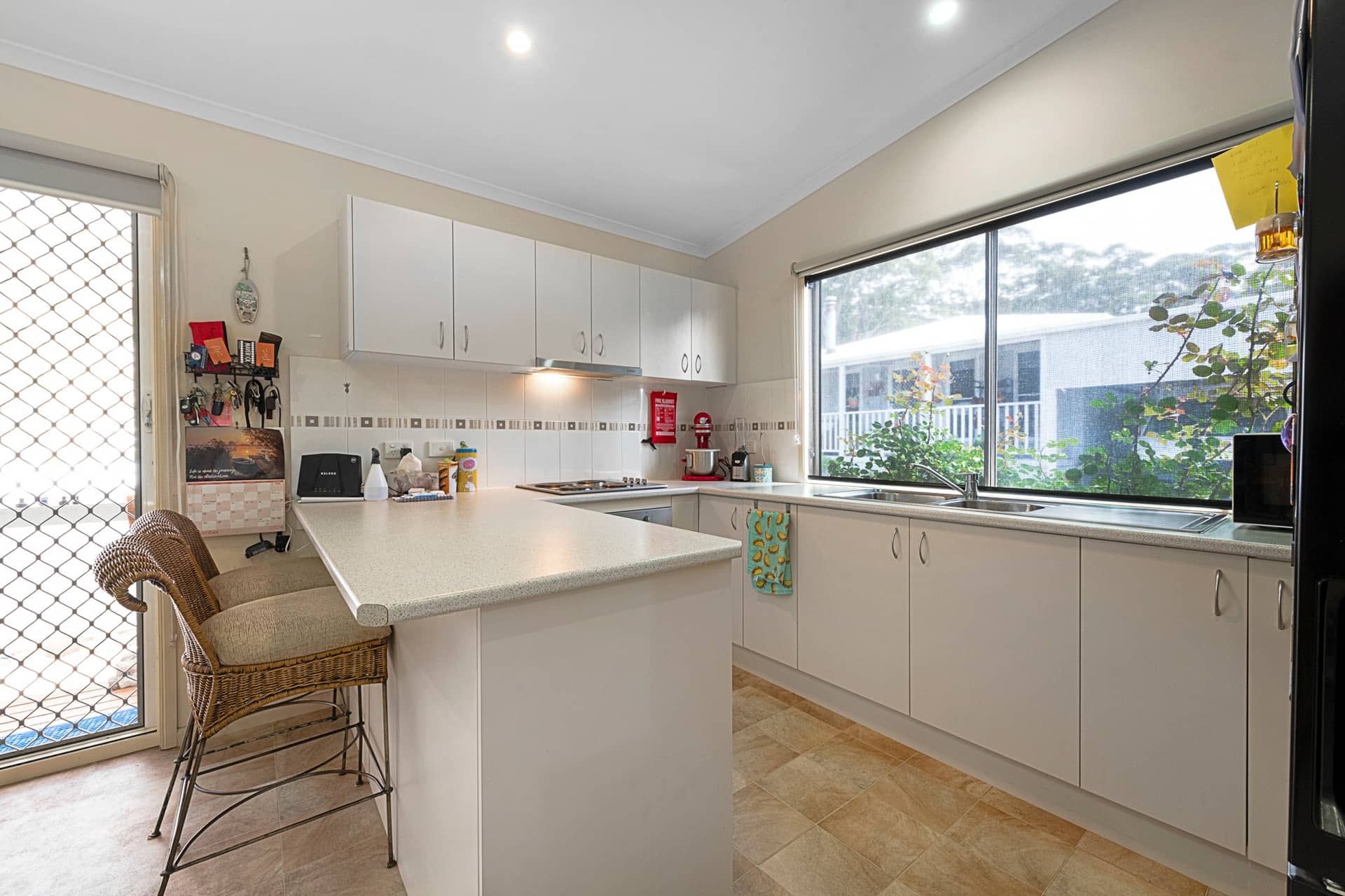 Light-toned kitchen with an L-shaped island bench, white cabinetry, and a window above the sink looking out to an Ingenia Lifestyle community.