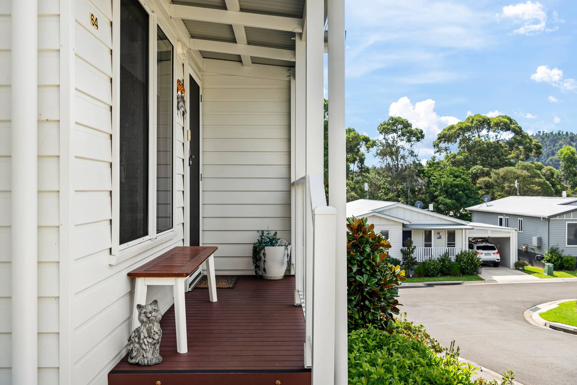Front porch of an Ingenia Lifestyle home with a wooden bench, cat statue, and plants. Community street view of homes and landscaping.