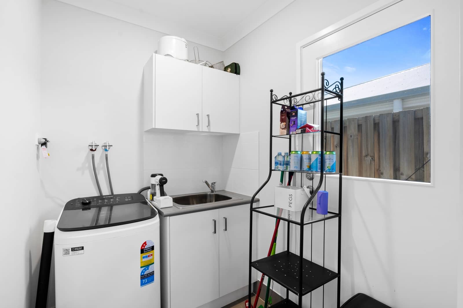 A laundry room with a washing machine, sink, and storage cabinets, featuring a window overlooking a fence and blue sky.