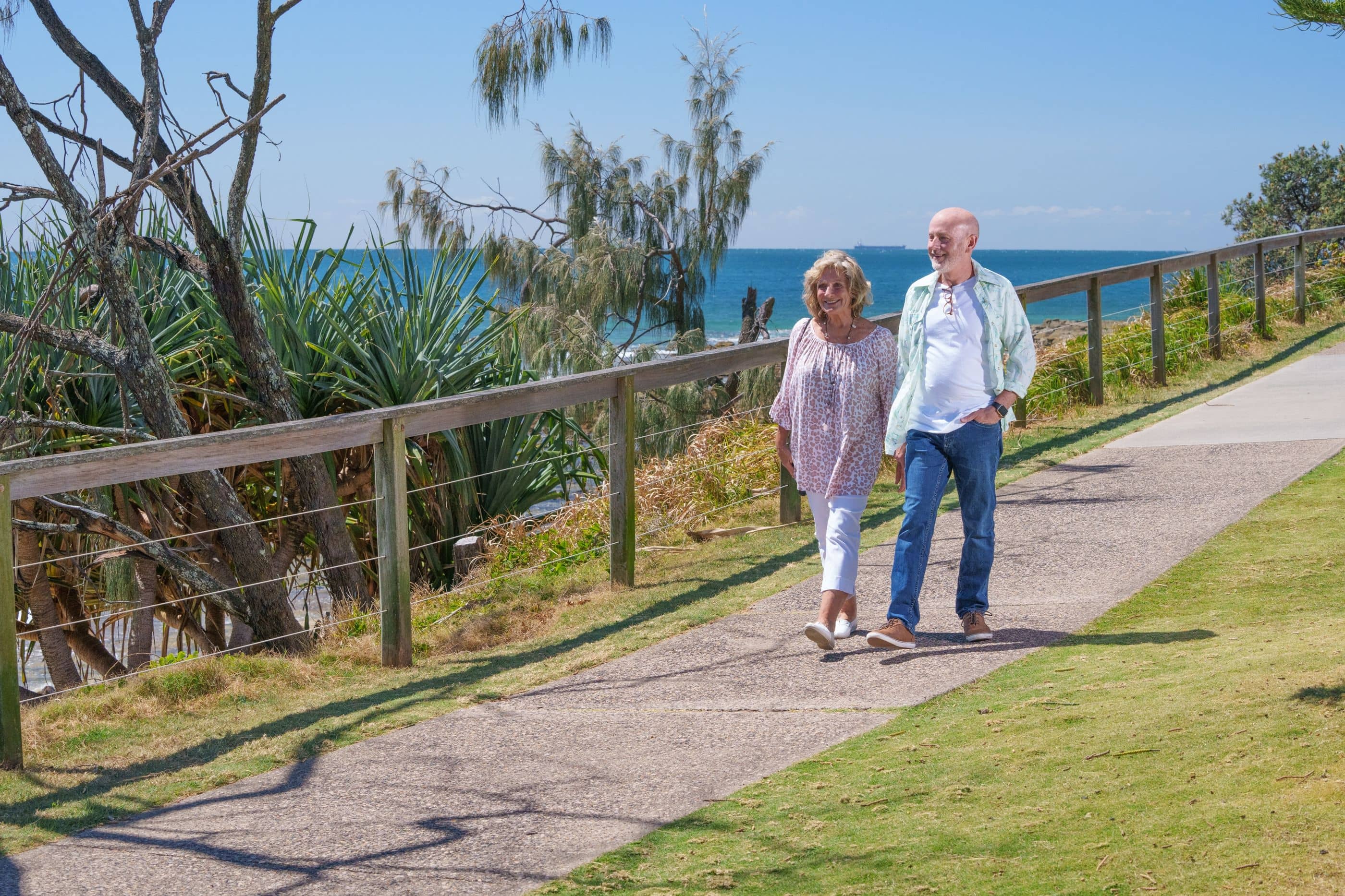 Two smiling people walk on a path overlooking the ocean. Coastal plants and a fence line the path in an Ingenia Lifestyle community.