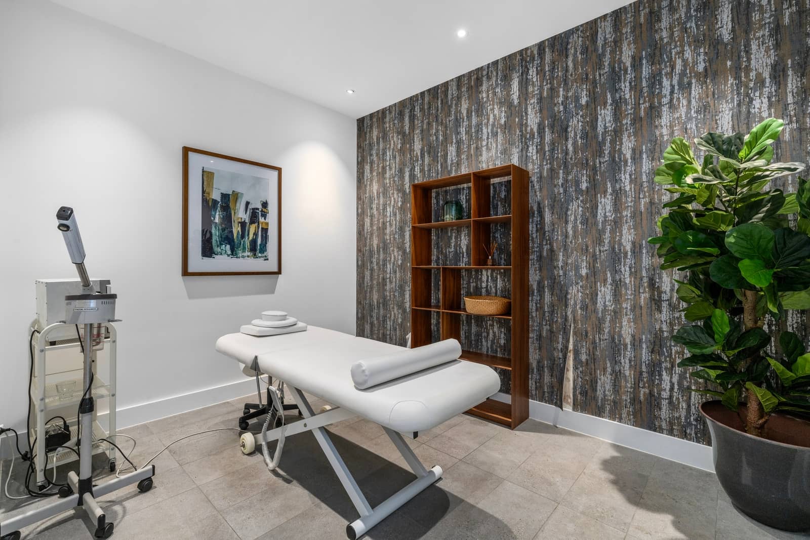 A modern treatment room in an Ingenia Lifestyle community with a white bed, facial steamer, and a dark textured accent wall.
