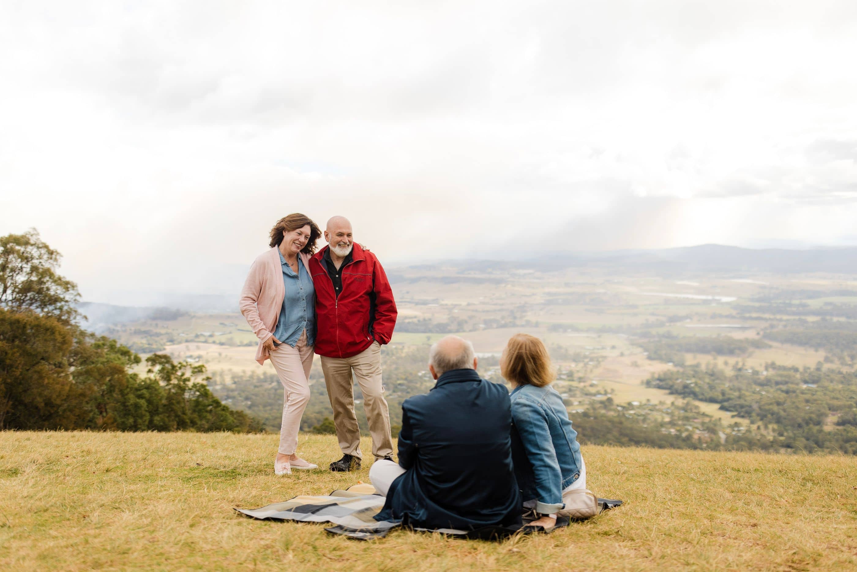 Four people on a grassy hill enjoying an outdoor gathering, overlooking a scenic valley. Ingenia Lifestyle community.