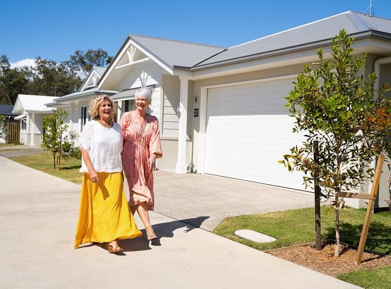 Two smiling women walk on a path past modern Ingenia Lifestyle homes with white garage doors.