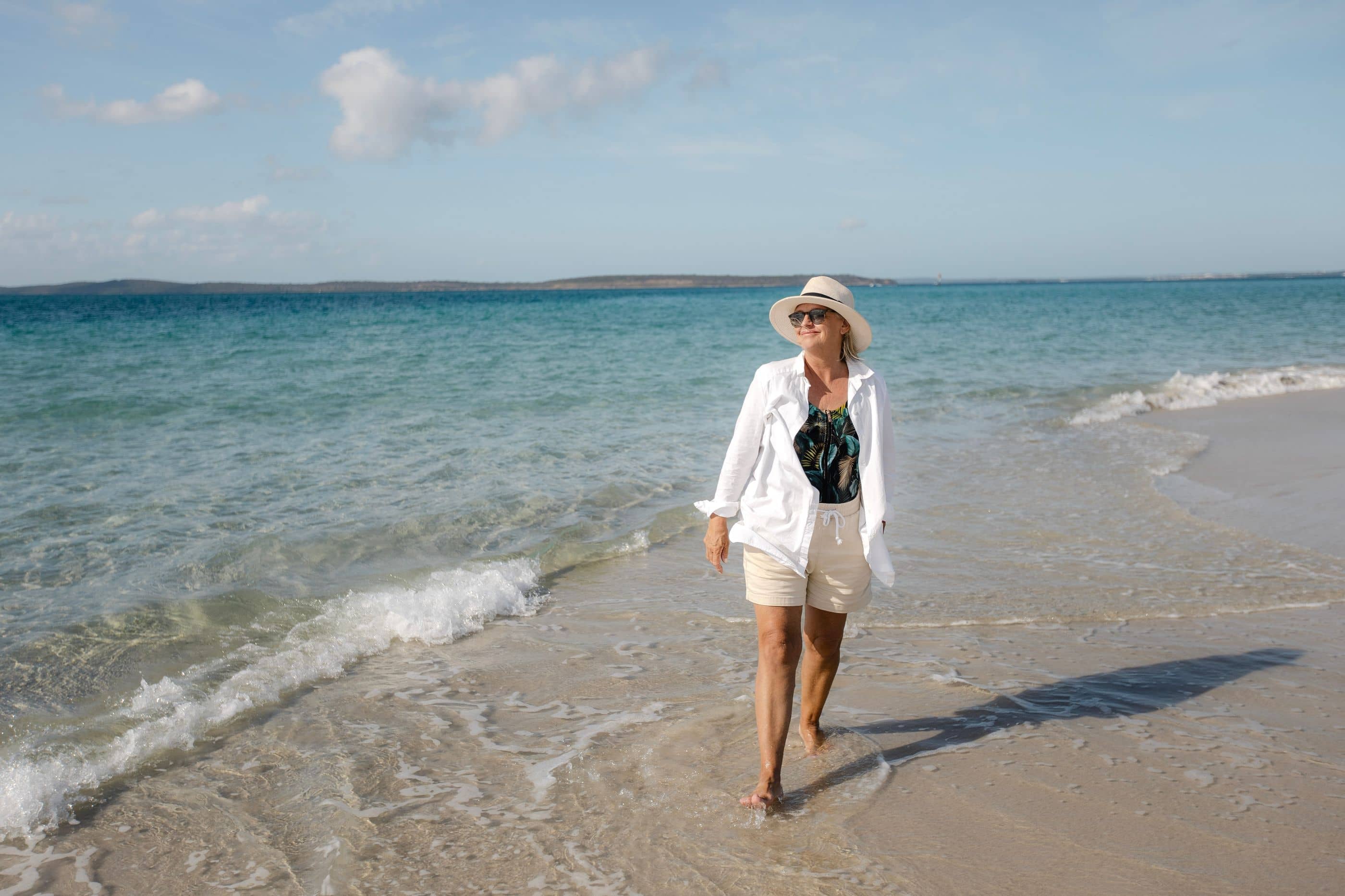 Woman in hat and sunglasses walks barefoot in the ocean waves on a sandy beach, showcasing an active lifestyle.