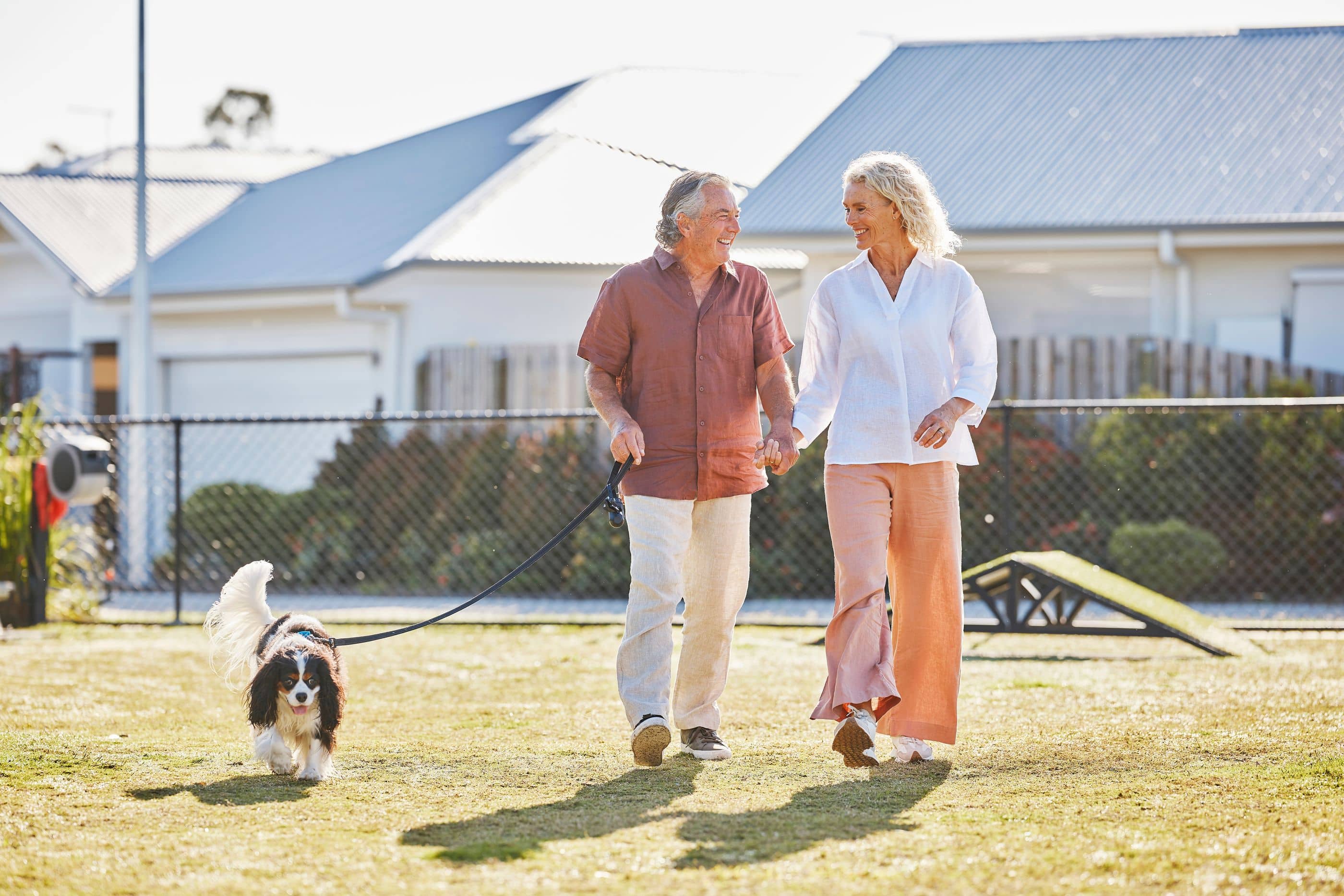 Two people holding hands taking a dog for a walk in a dog park.