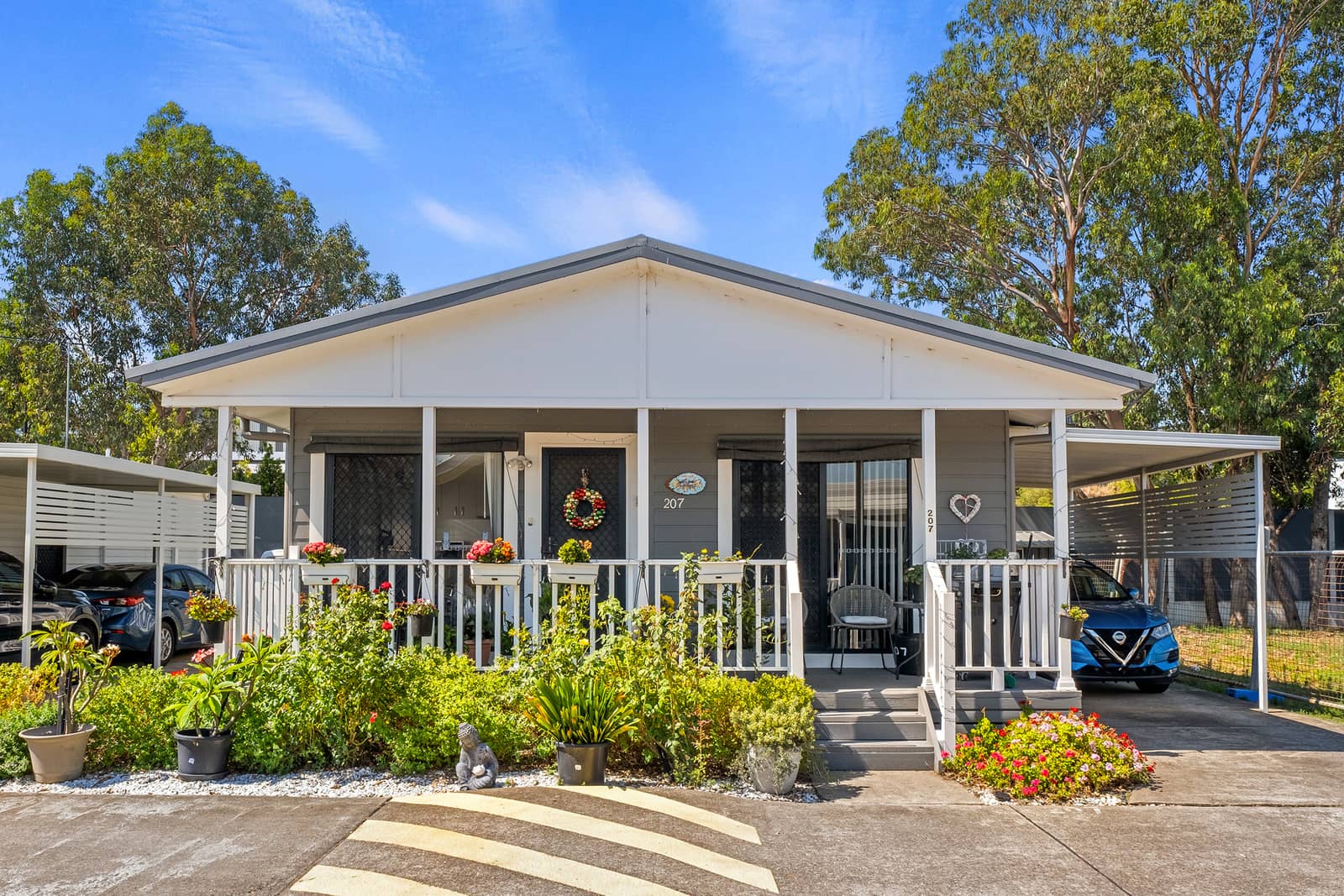 A grey land lease home with a white gabled roof and a covered verandah, surrounded by lush gardens.
