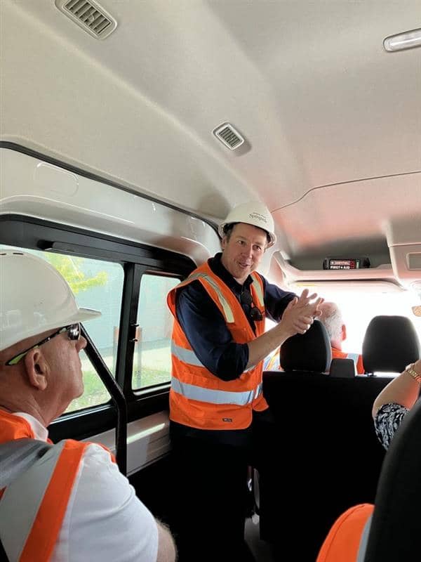 Man in a hard hat and high-visibility vest speaks to people in a van, possibly on a community tour.