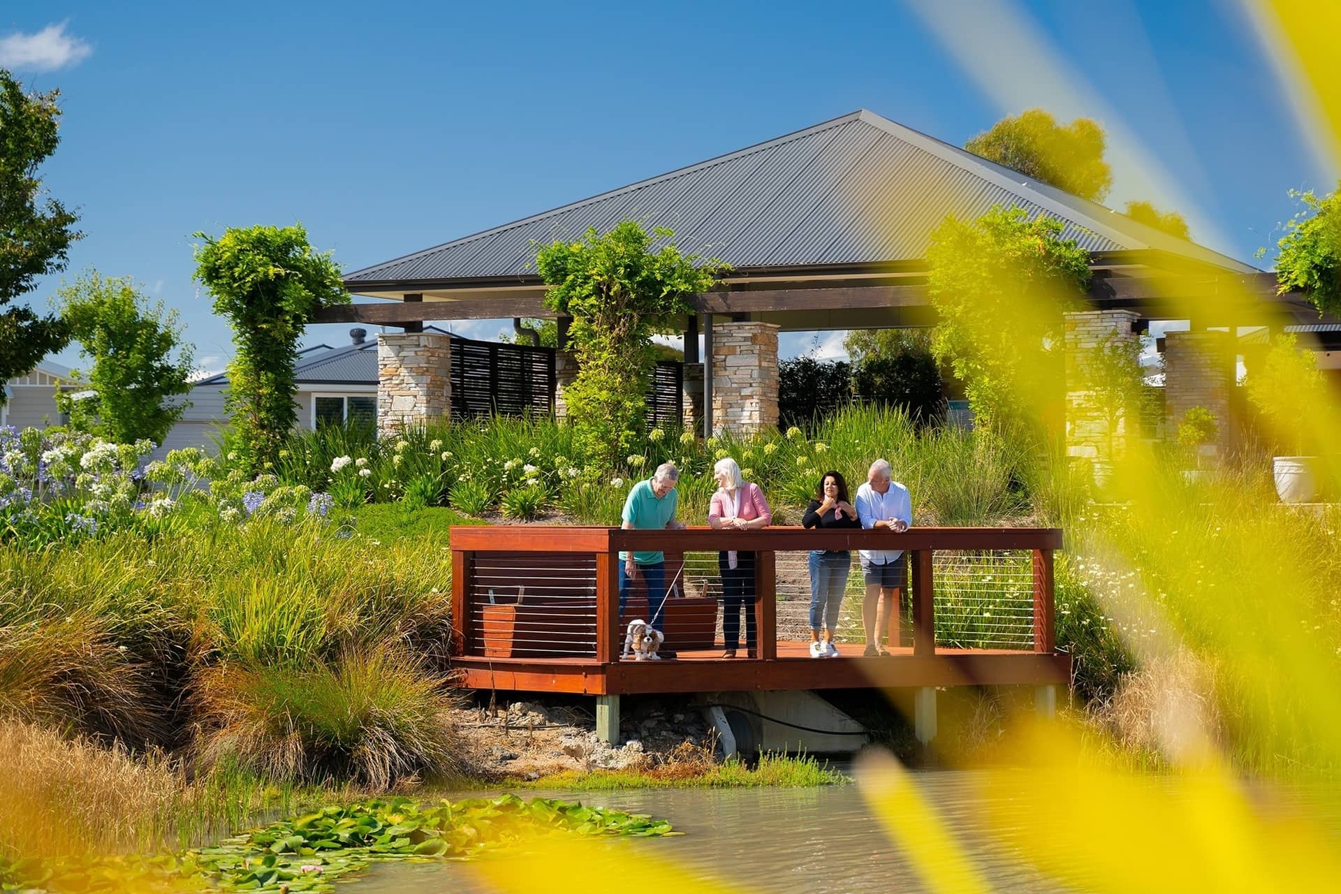 Four people and a small dog stand on a wooden deck overlooking a pond and landscaped grounds of an Ingenia Lifestyle community.