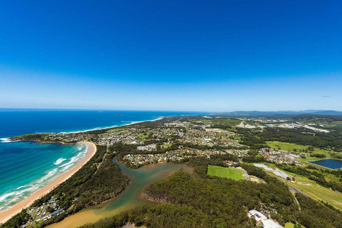 Aerial view of a coastal town featuring a long beach, ocean, river, residential area, and green landscape.