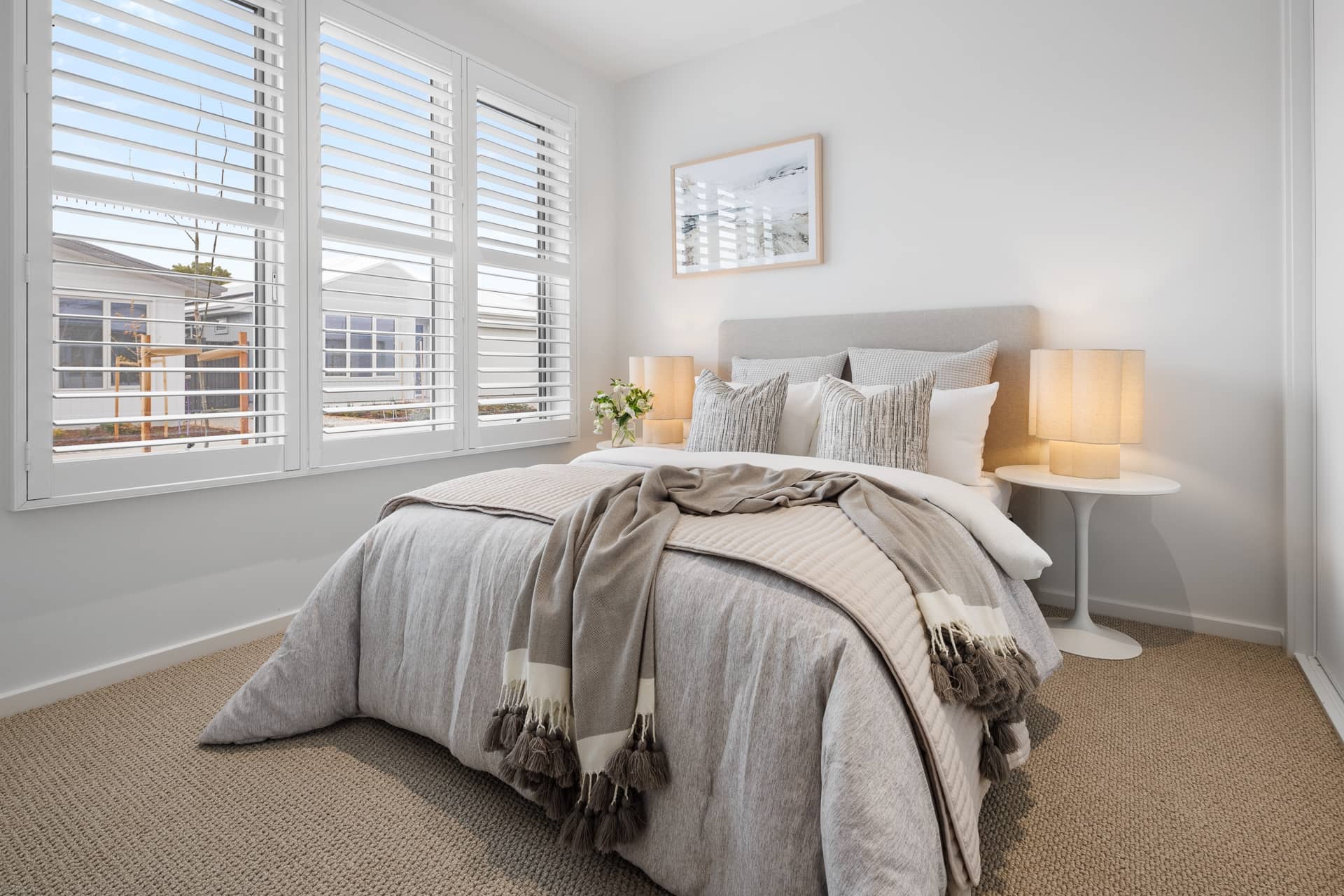 A bedroom in an Ingenia Lifestyle home with a bed, two bedside tables, lamps, and white plantation shutters on large windows.