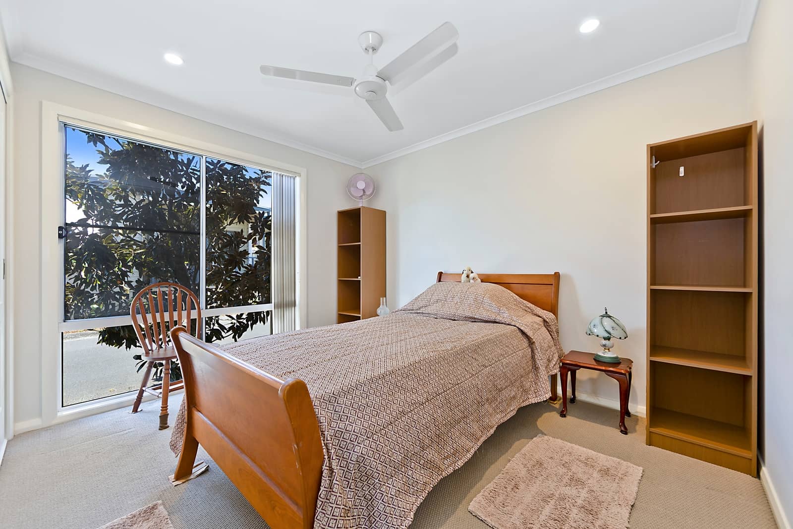 Bedroom with wooden bed, patterned quilt, window view of trees, ceiling fan, and wooden shelves in an Ingenia Lifestyle home.
