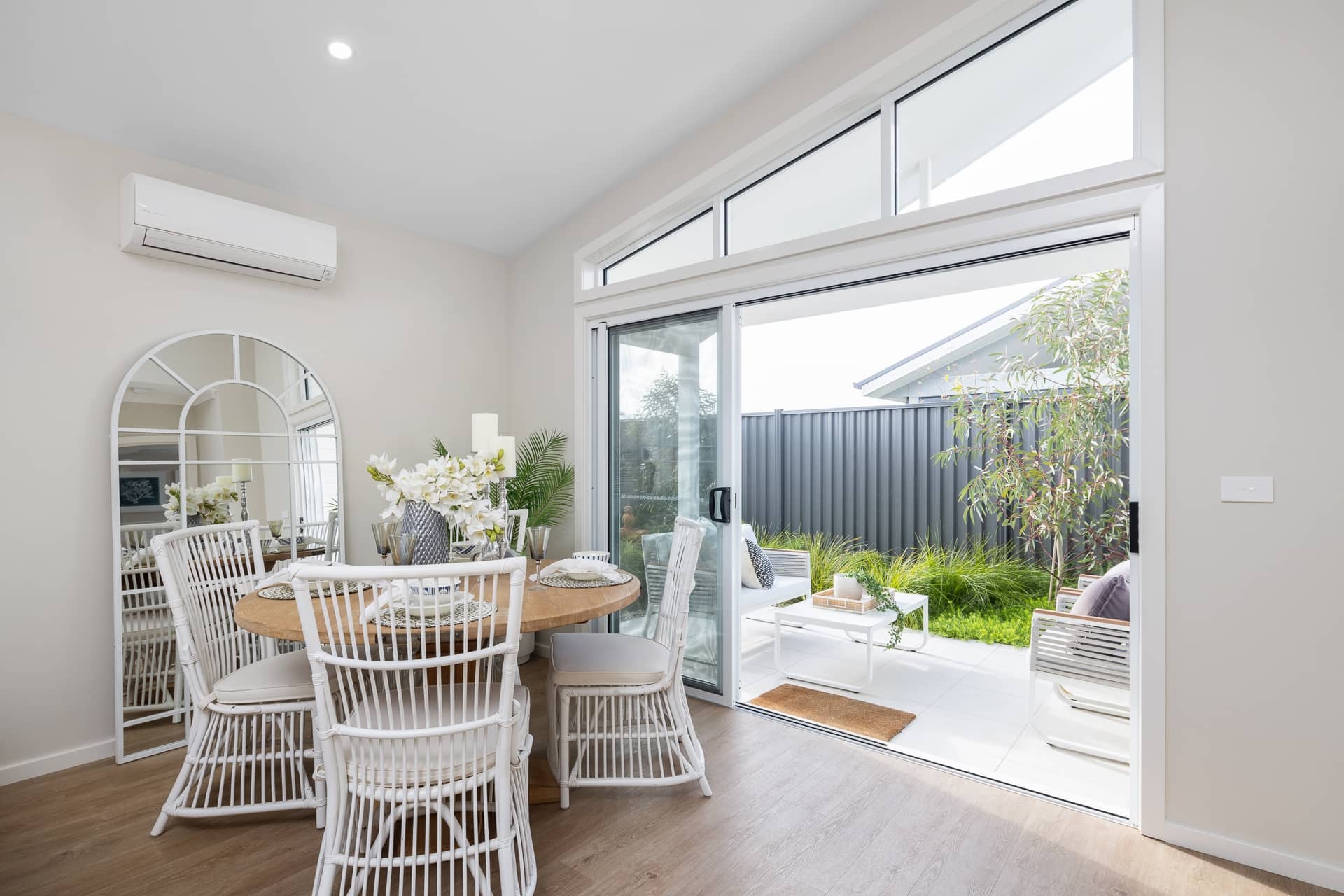 Modern dining area with a round wooden table and white rattan chairs, opening to a paved outdoor patio.