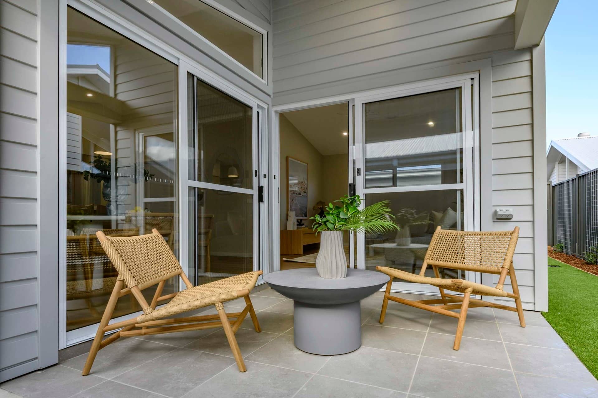 Two woven chairs flank a grey round table with a vase of greenery on a tiled patio.