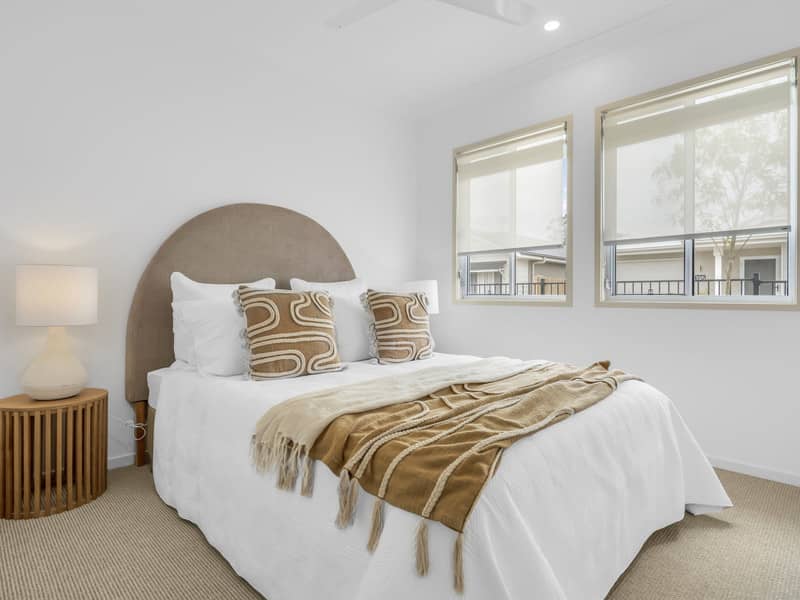 Bedroom with a queen bed, neutral tones, and two windows overlooking an Ingenia Lifestyle community.