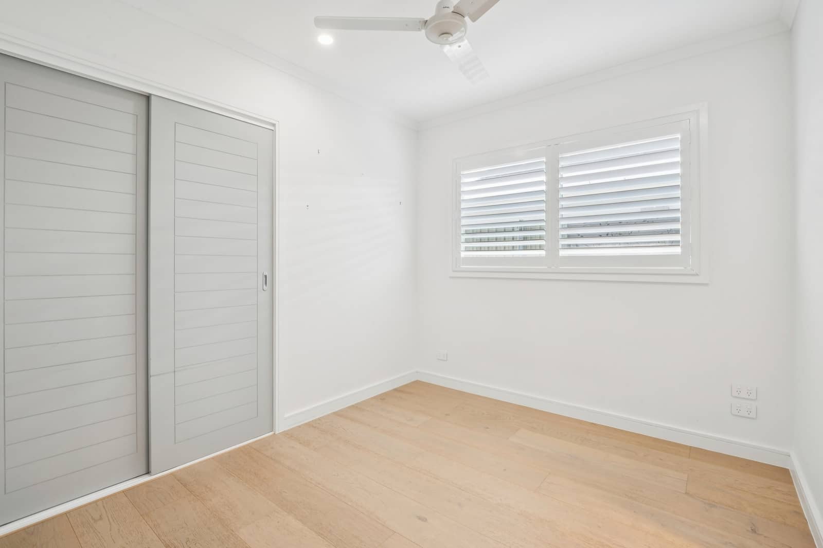 Interior view of a bright, empty bedroom with sliding doors, window shutters, and light wood floor.