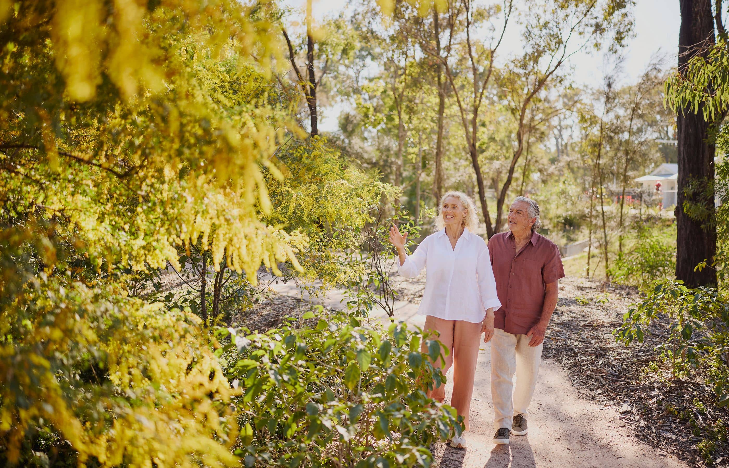 Two people walk on a dirt path through a lush, green, and yellow landscape, with trees and foliage surrounding them.