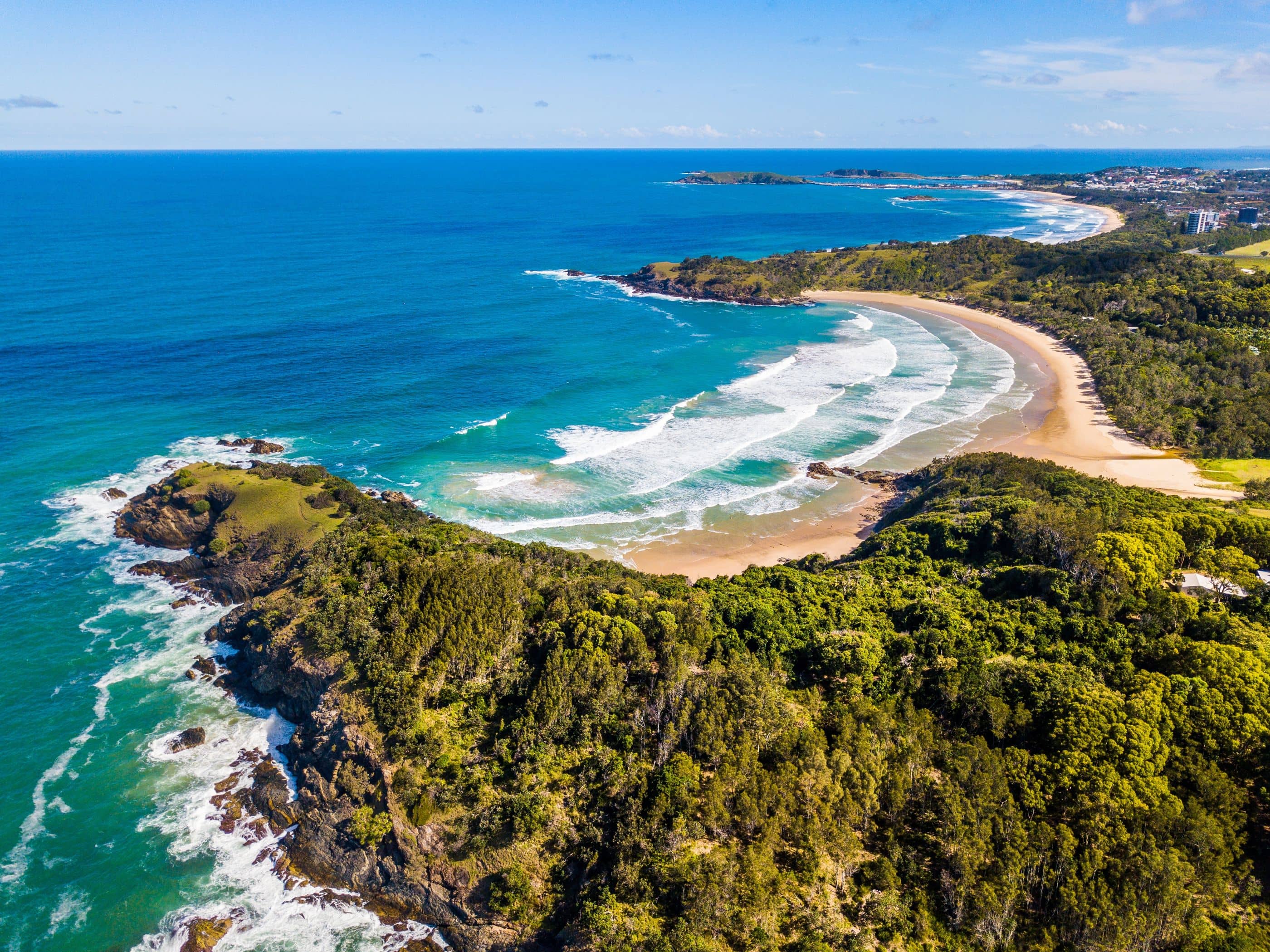 Aerial view of a coastal landscape with rocky outcrops, a sandy beach, and green, treed hills.