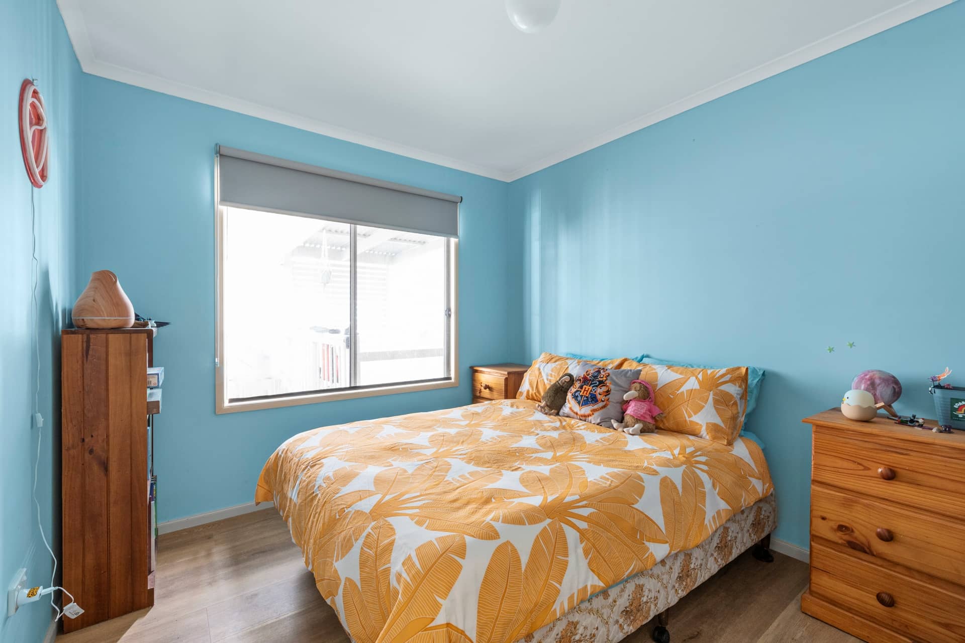 Bedroom in an Ingenia Lifestyle home: light blue walls, wood floor, bed with tropical orange and white bedding, and window.
