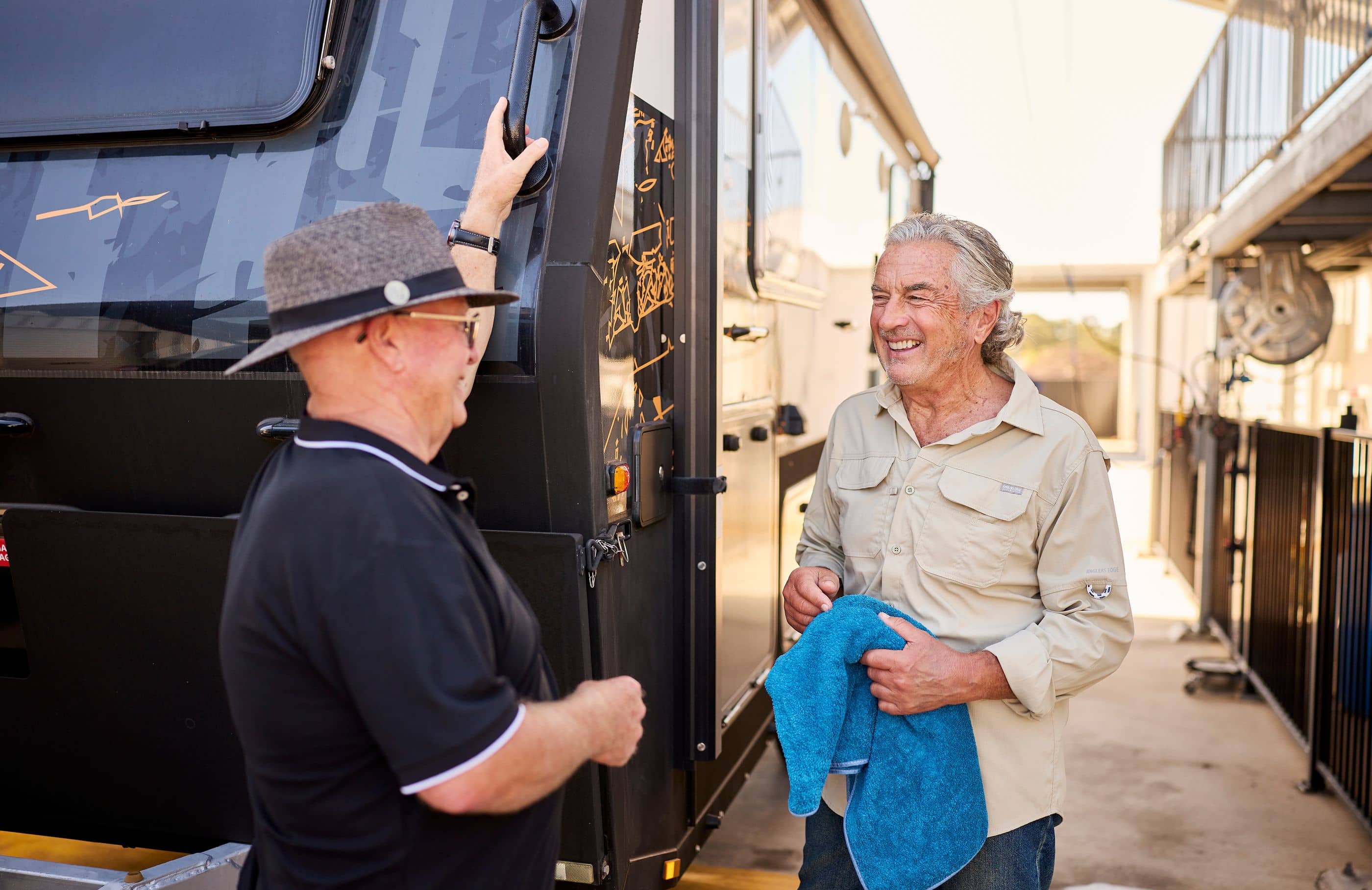 Two men talking next to a large caravan or RV. One man wears a grey hat and black shirt, reaching for the caravan. The other man wears a beige shirt and holds a blue towel.
