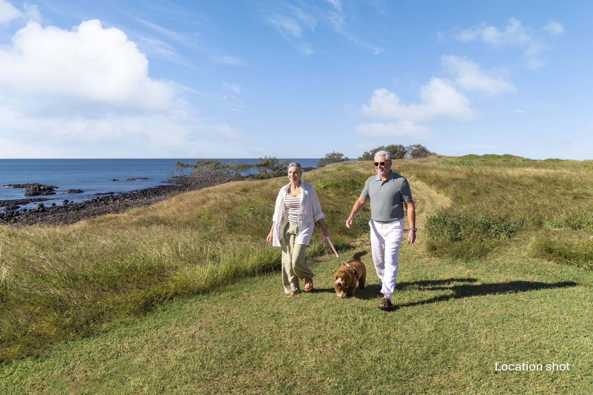 A couple walks a brown dog on a grassy path with the ocean and rocky shoreline in the background.