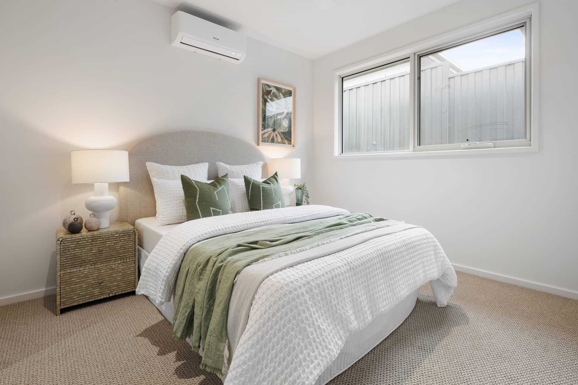Bedroom in an Ingenia Lifestyle home featuring a bed with white and green linen, bedside lamps, a window, and air conditioning.