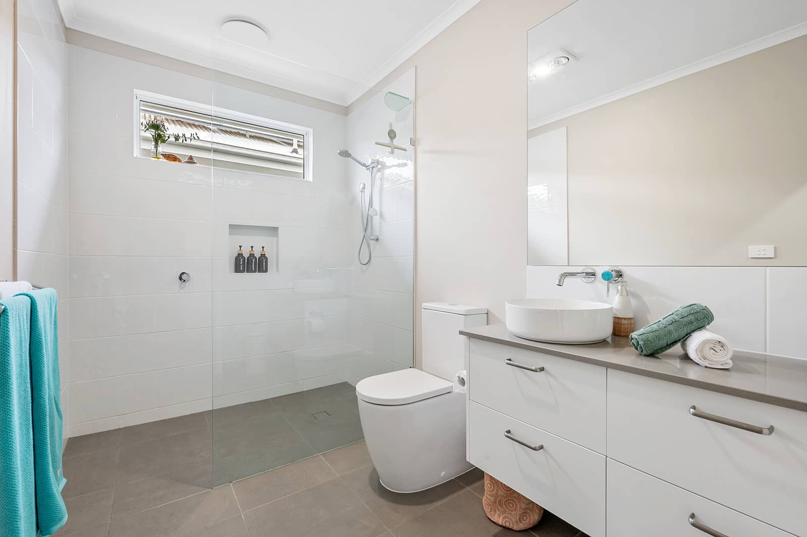 Modern bathroom with white tiled walls, walk-in shower, white vanity, basin, toilet, and grey tiled floor.