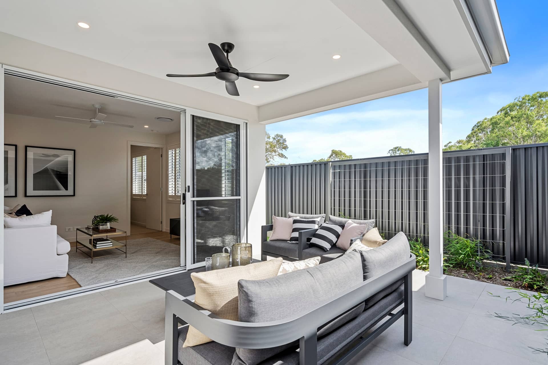 Covered outdoor patio with outdoor lounge seating and a ceiling fan, overlooking a backyard fence.