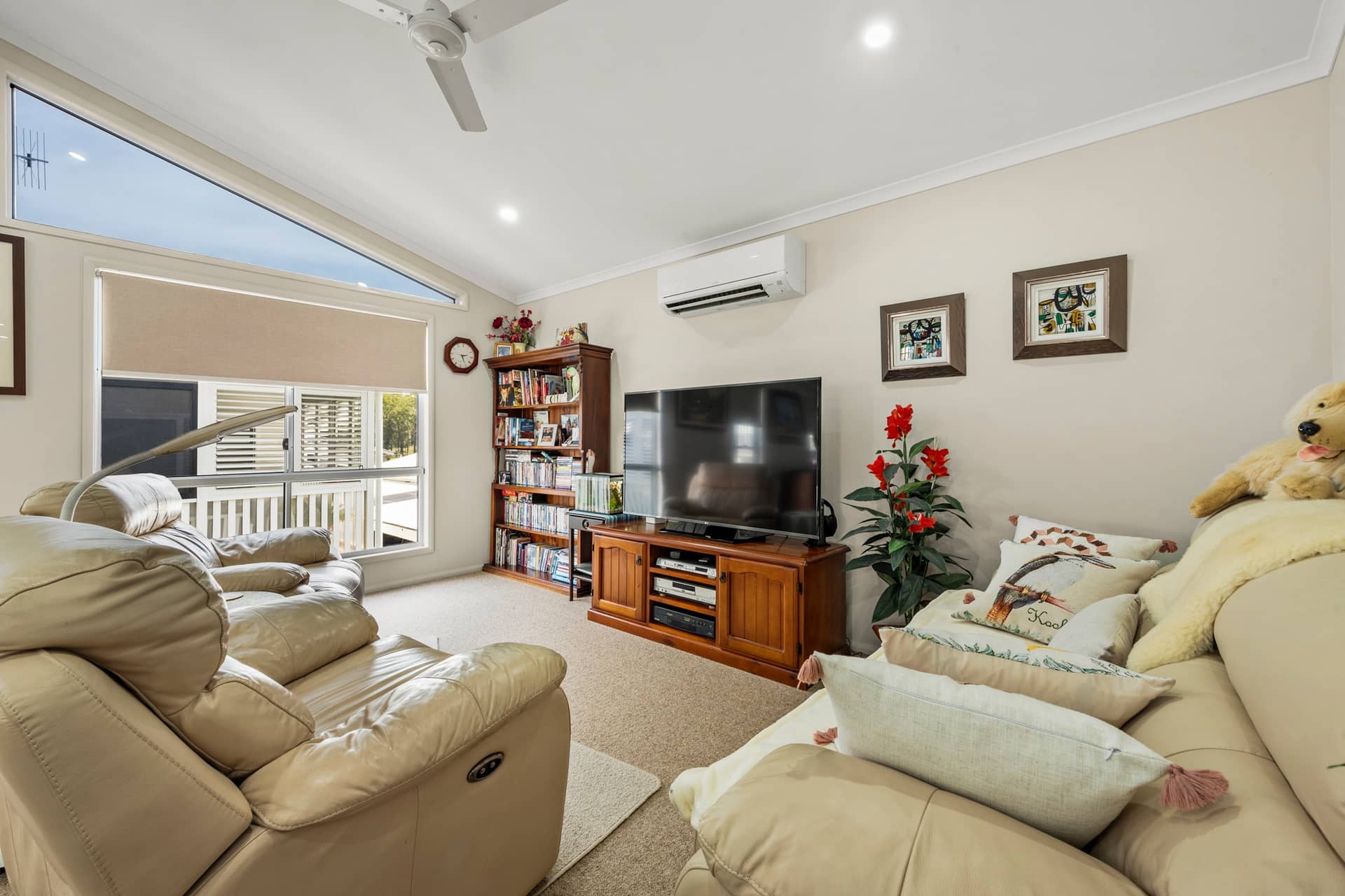 A light-filled living room inside an Ingenia Lifestyle home, with beige sofas, a TV, bookshelf, large windows, ceiling fan, and air conditioning.