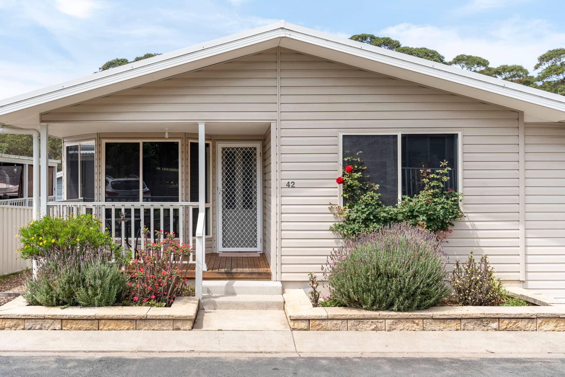 Exterior of an Ingenia Lifestyle home with beige siding, a front porch, and landscaped garden beds with flowers.