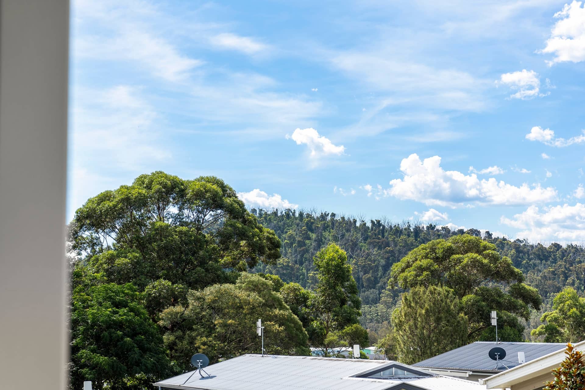 View from an Ingenia Lifestyle home, showing grey rooftops, green trees, and a distant forested hill under a blue, cloudy sky.