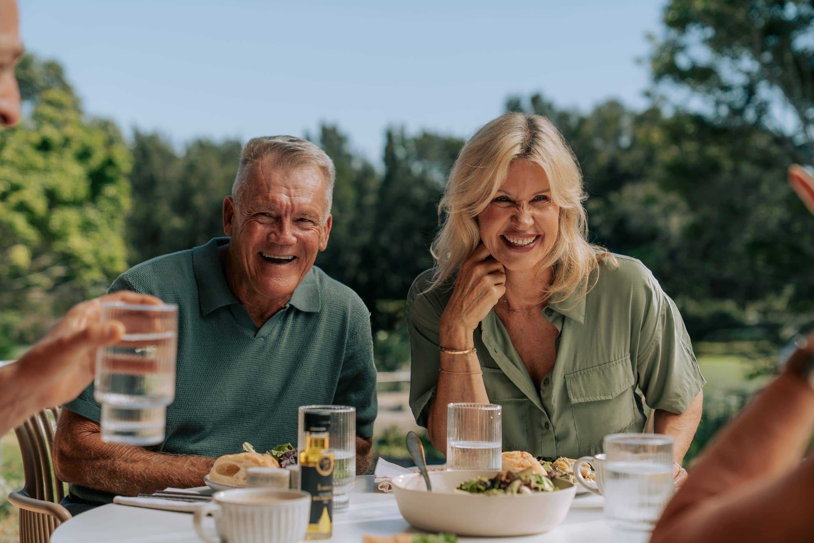 Two laughing residents enjoy an outdoor meal at an Ingenia Lifestyle community, with green trees behind.