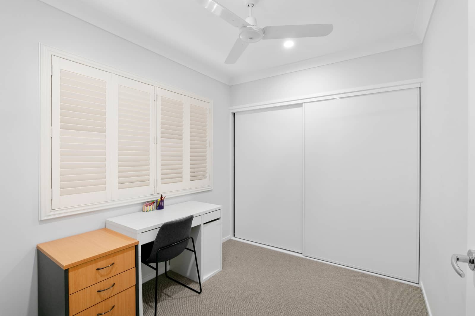A white desk and chair are in front of plantation shutters in a room with a ceiling fan and sliding wardrobe doors.