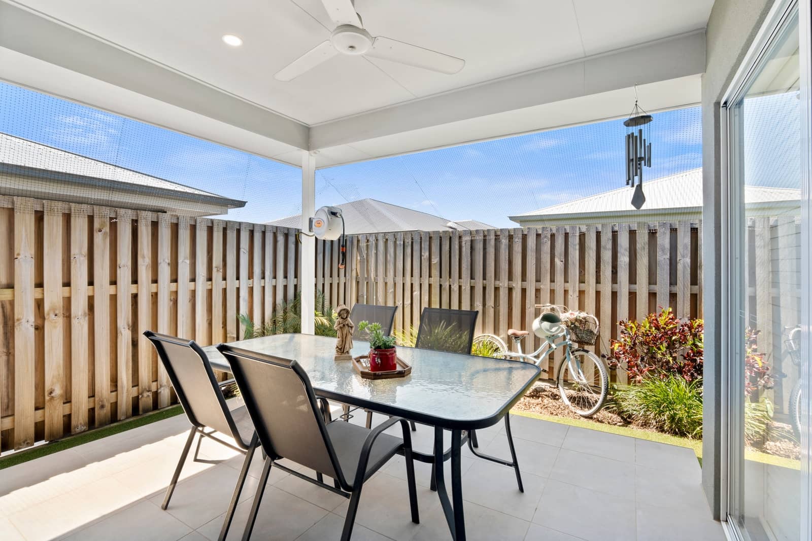 An outdoor patio with a glass-top table and four chairs overlooks a wooden fence and a light blue bicycle.