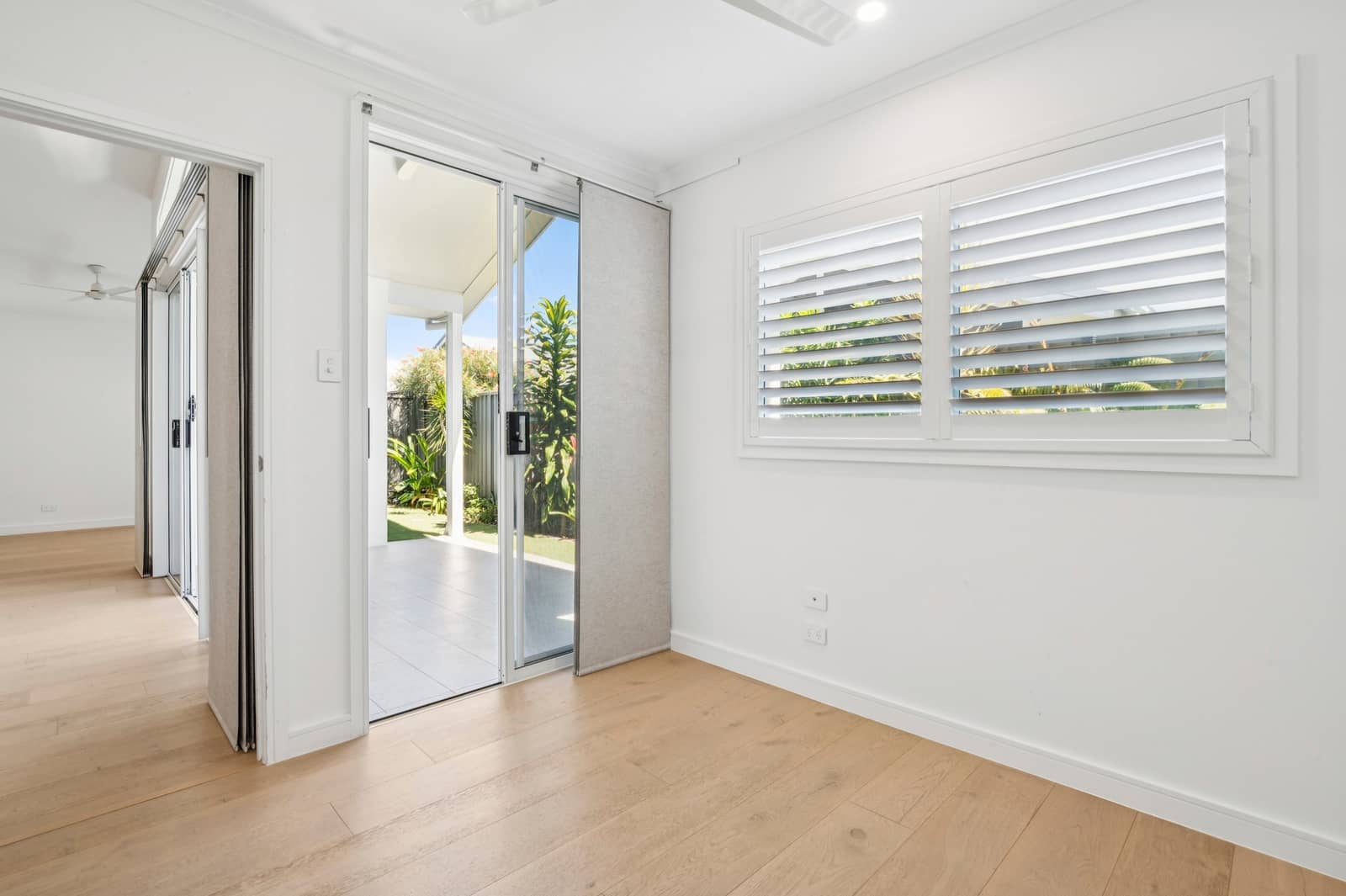 An empty room with light wood floors, sliding glass doors, and plantation shutters.