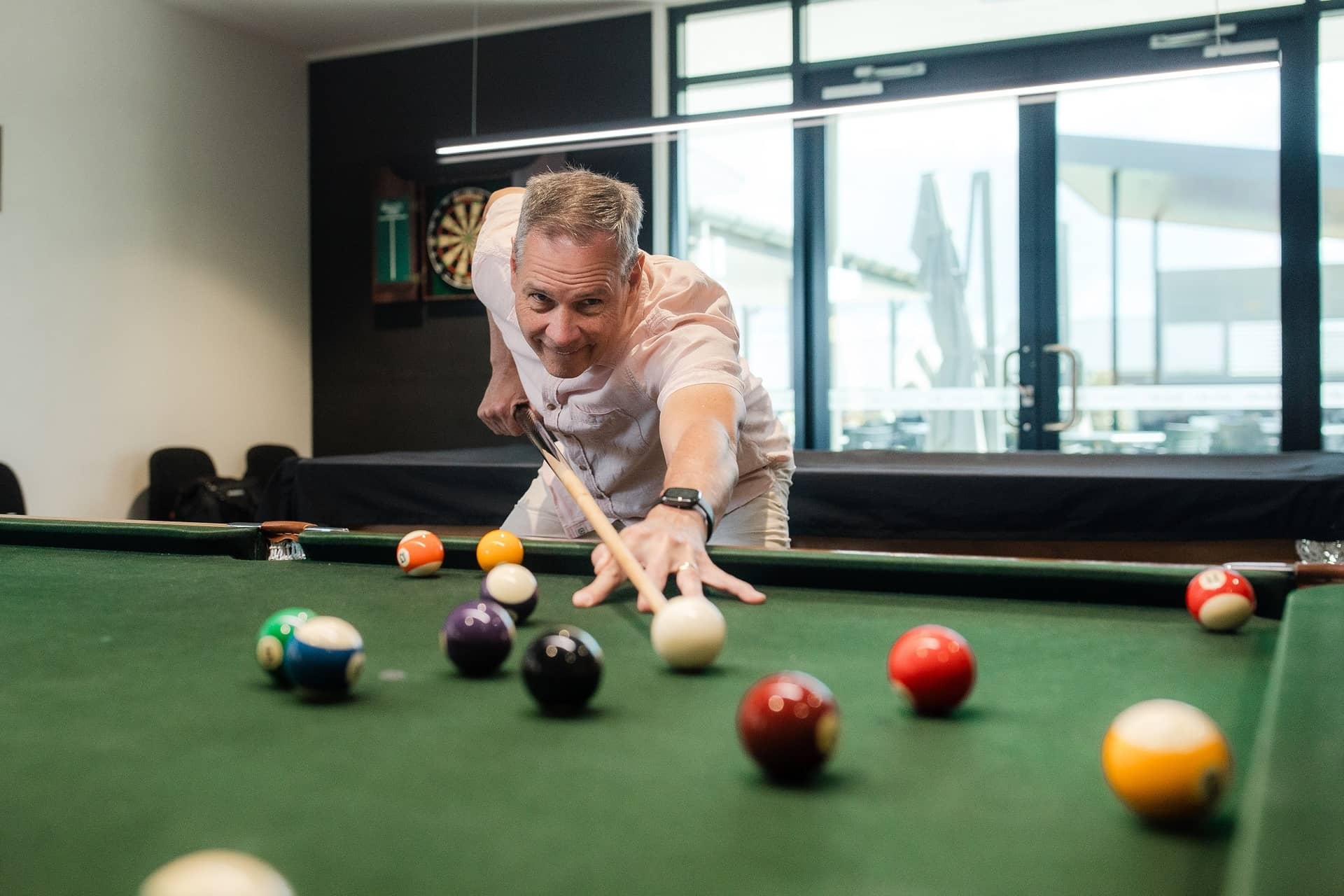 A smiling man leaning over a green pool table, aiming a cue stick at billiard balls in an Ingenia Lifestyle community facility.